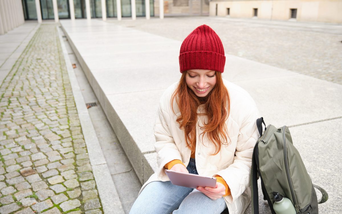 Beautiful redhead woman in red hat, sits with backpack and thermos, using digital tablet outdoors, connects to wifi, texts message, books tickets online.
